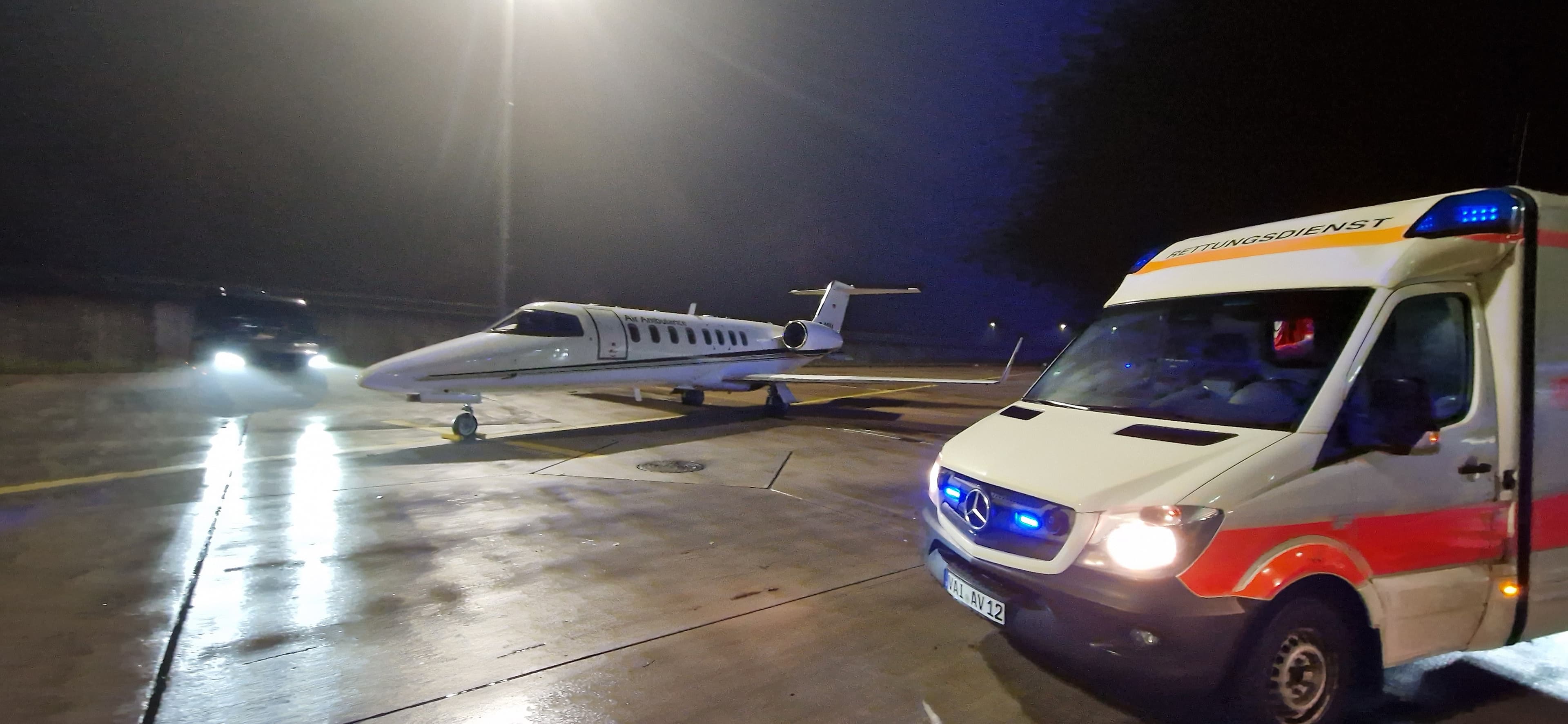 Ambulance with blue lights next to a private jet on a wet runway at night.