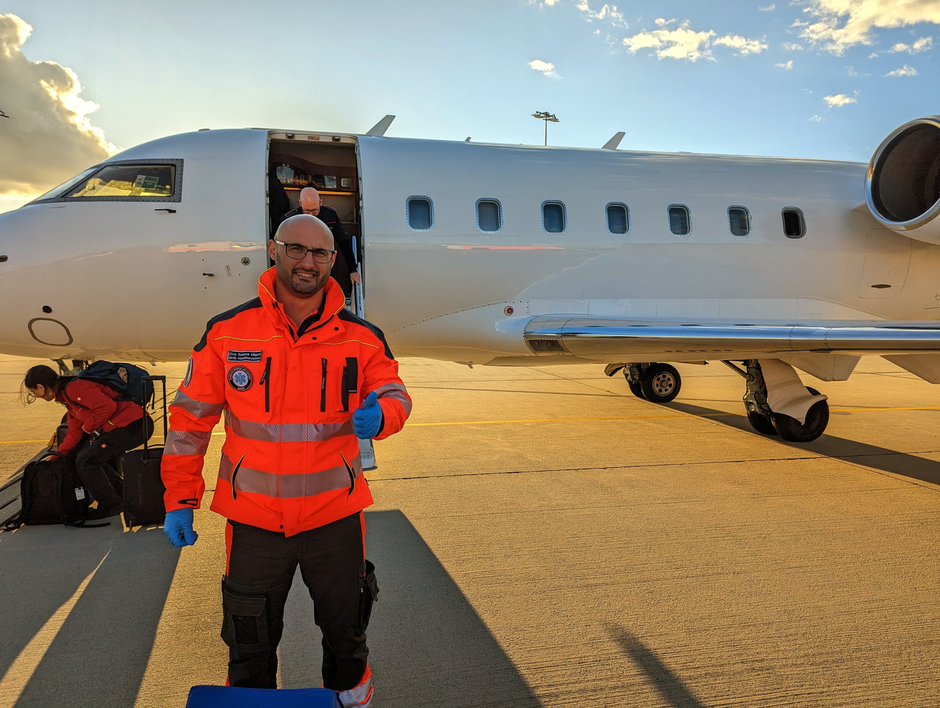 Paramedic in orange jacket giving a thumbs up beside a white private jet on tarmac.