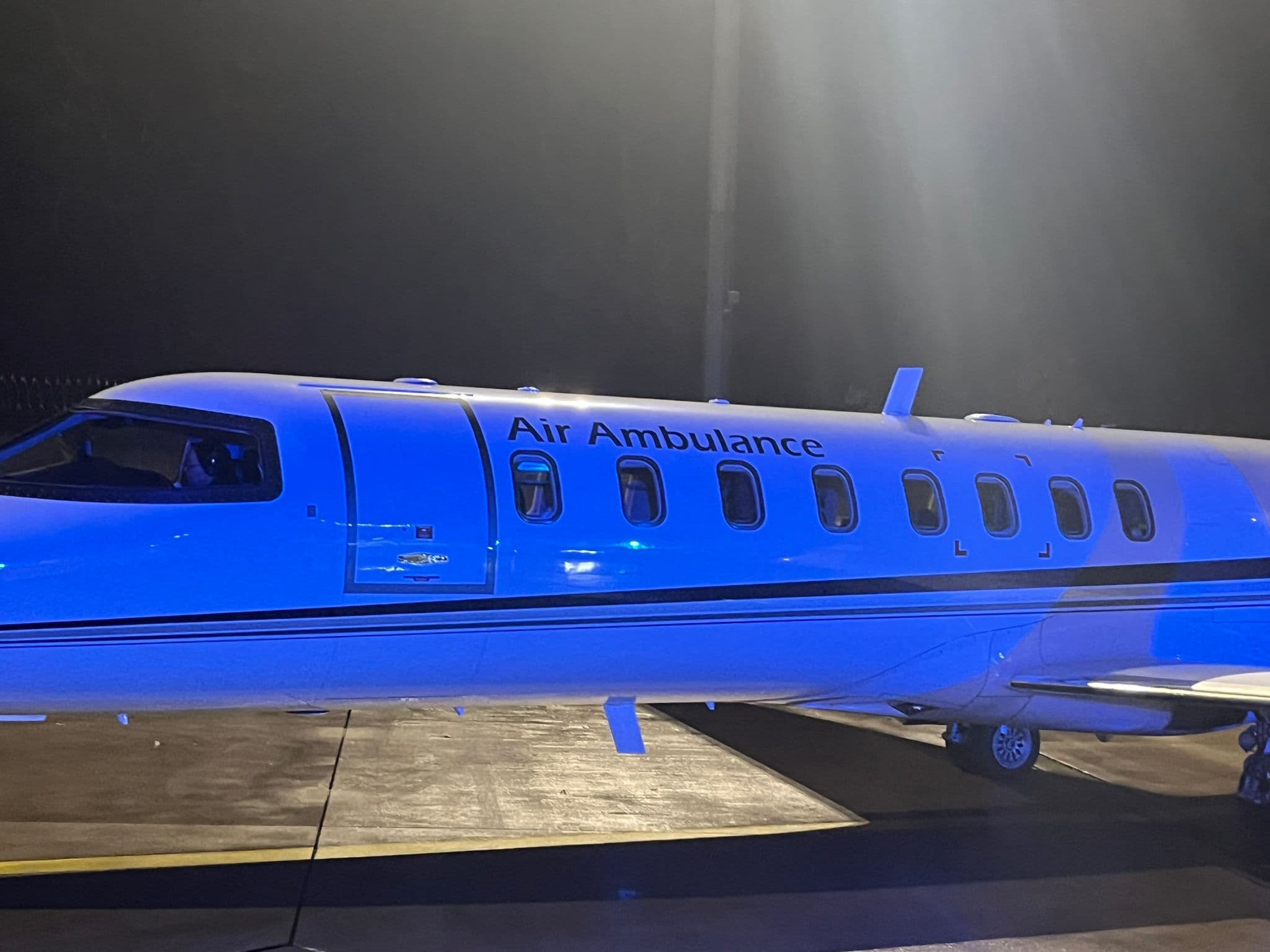 White air ambulance jet parked on a tarmac at night under bright blue lighting.