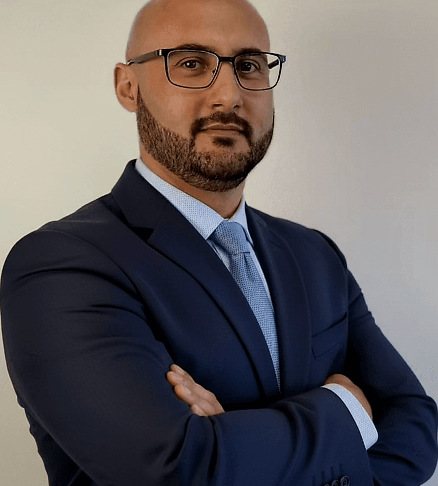 Bald man with glasses and beard wearing a navy suit and tie, arms crossed.